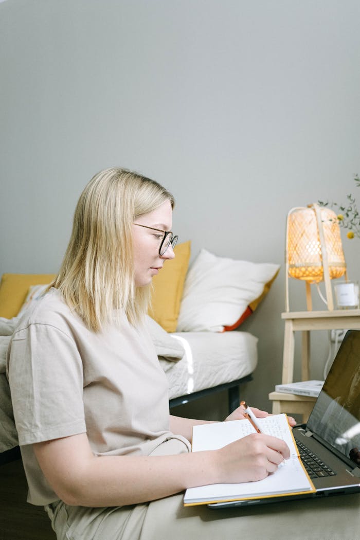 A woman focusing on work with a laptop and notebook in a cozy bedroom.